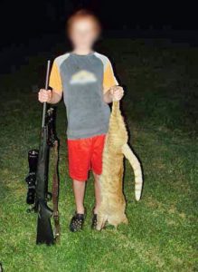 Boy holds dead cat in left hand and rifle in the right